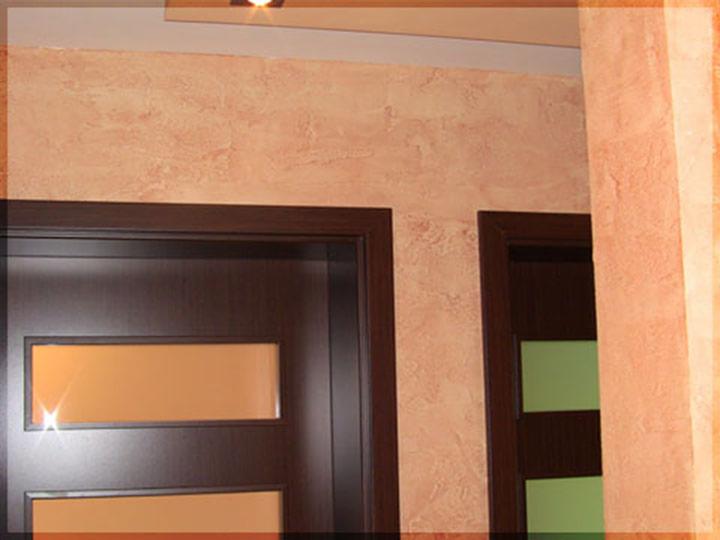 Wykończenie ścian w Krakowie: Dekoracyjna gładź w odcieniach beżu i brązu, imitująca naturalny kamień. Widoczne fragmenty dwóch ciemnych drzwi z przeszkleniami.