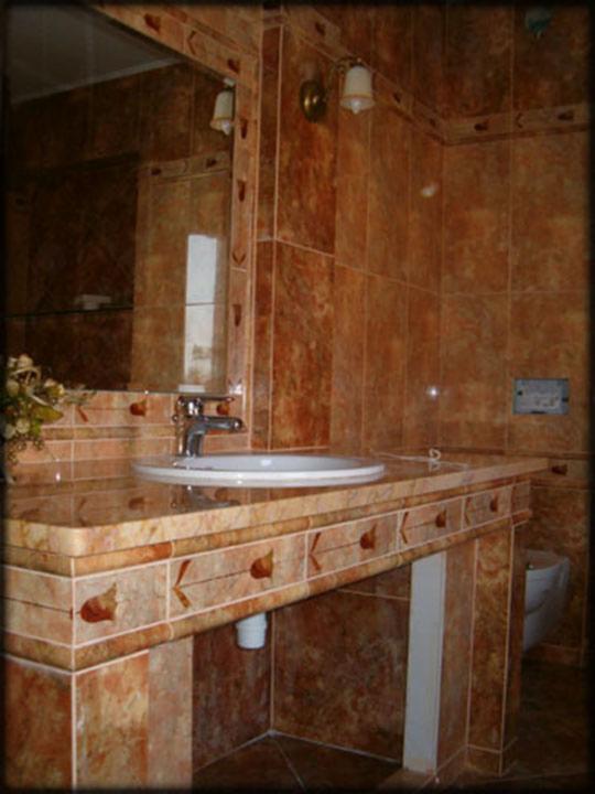 Wykończona łazienka z brązowymi, wzorzystymi płytkami na ścianach i zabudowie umywalki. Widoczna umywalka, lustro i lampki. Całość w ciepłej tonacji.