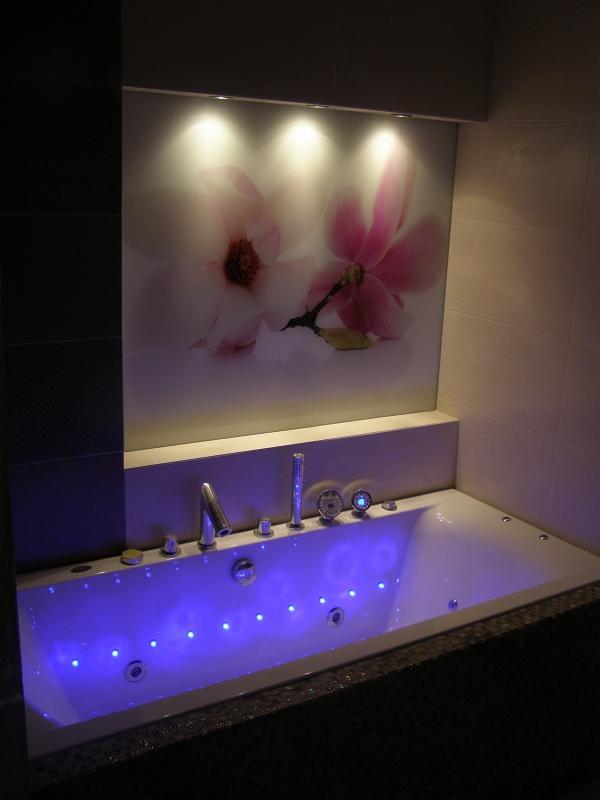 Nowoczesna wanna z hydromasażem i podświetleniem LED w łazience wykończonej płytkami. Nad wanną panel dekoracyjny z motywem kwiatowym i oświetleniem punktowym.