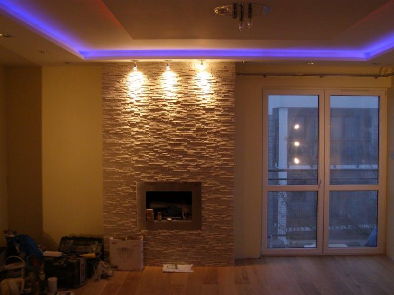 Salon z kominkiem obłożonym dekoracyjną, kamienną płytką w odcieniach beżu, oświetlonym trzema punktowymi lampami. Sufit z niebieskim oświetleniem LED.