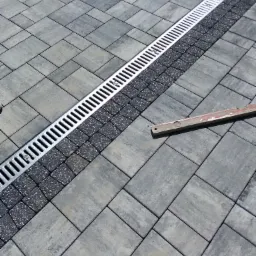 Szara kostka brukowa ułożona we wzór jodełki z liniowym odpływem wody i poziomicą leżącą na powierzchni.