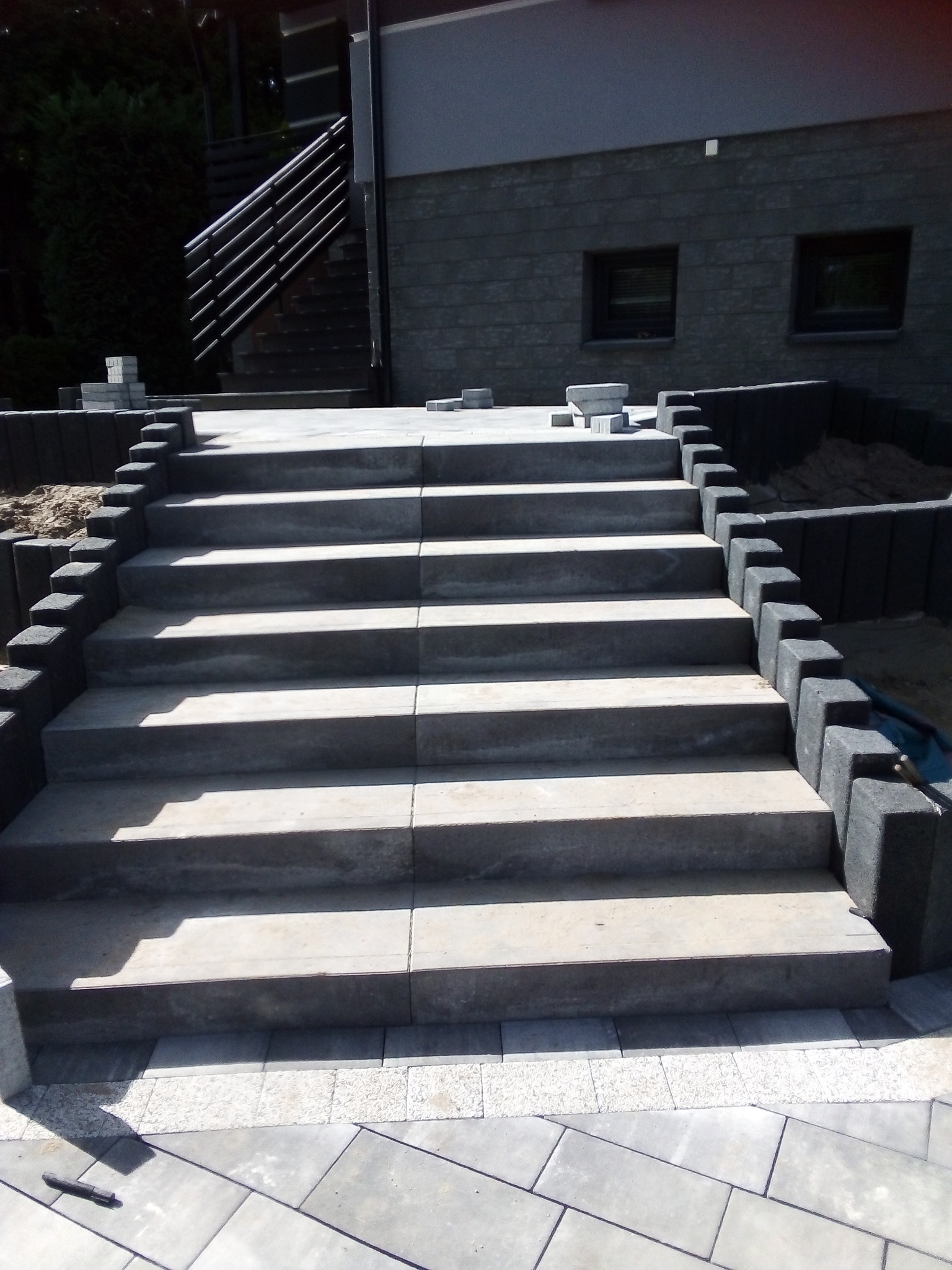 Schody zewnętrzne z szarej kostki brukowej, obrzeża z ciemnych betonowych elementów, w tle budynek z szarej cegły i metalowe schody.