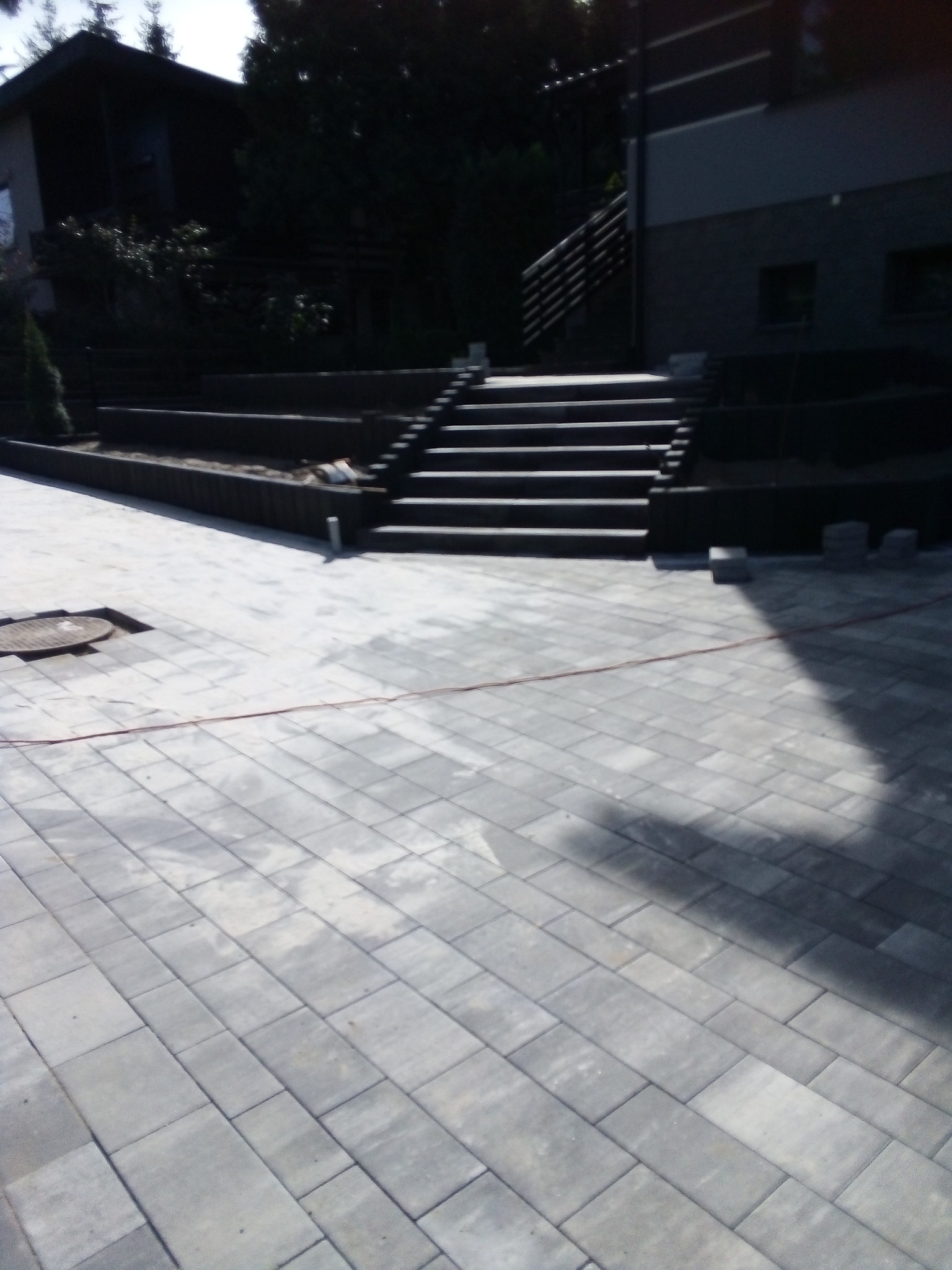 Ułożona szara kostka brukowa na podjeździe, z widocznymi schodami prowadzącymi do domu i niezakończonymi elementami obrzeży.