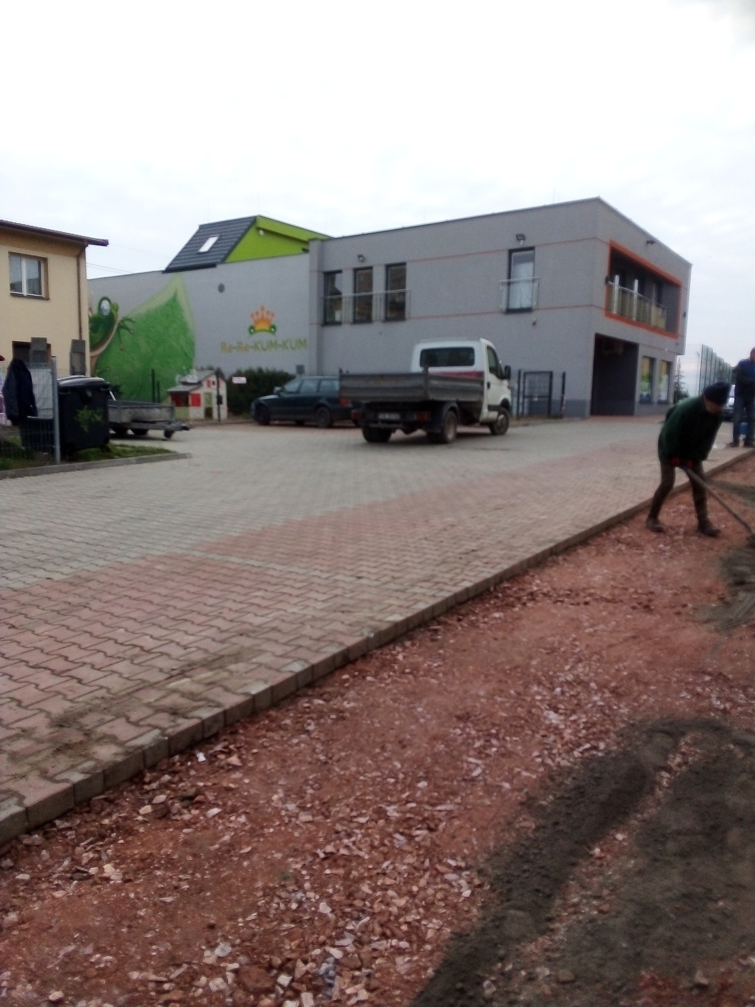 Przygotowanie podłoża pod kostkę brukową na terenie przedszkola, widoczny pracownik wyrównujący teren, obok ułożona kostka brukowa i zaparkowany samochód dostawczy.