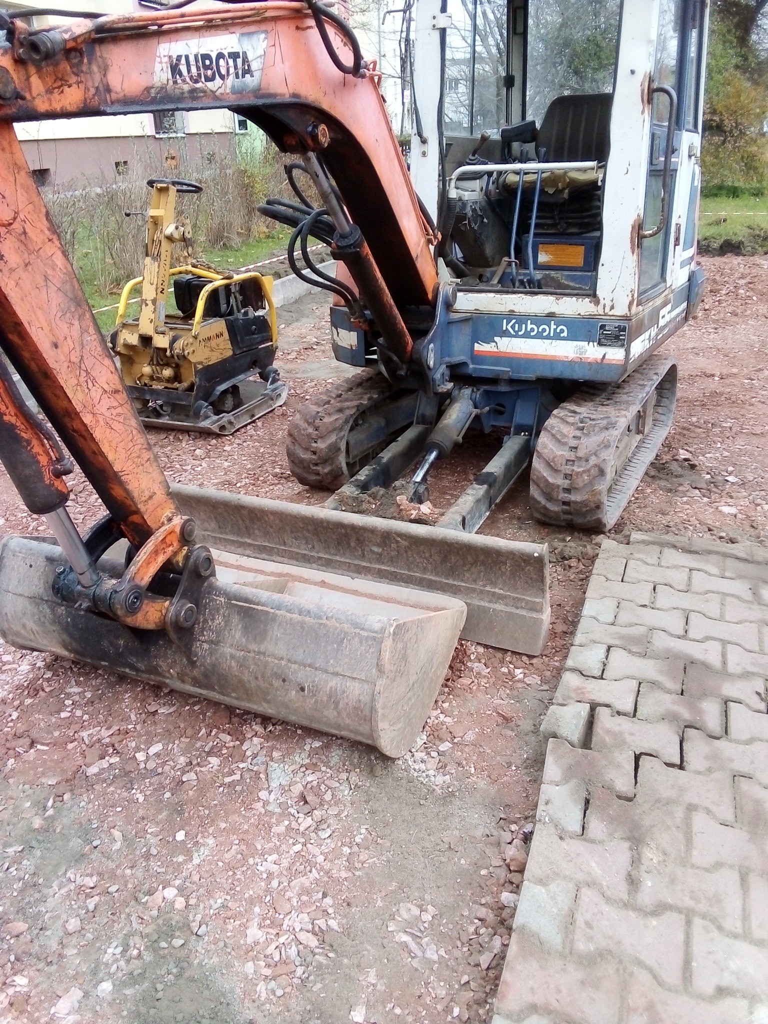 Pomarańczowo-biała mini koparka Kubota z łyżką na żwirowym podłożu, obok zagęszczarka gruntu i ułożona częściowo kostka brukowa.