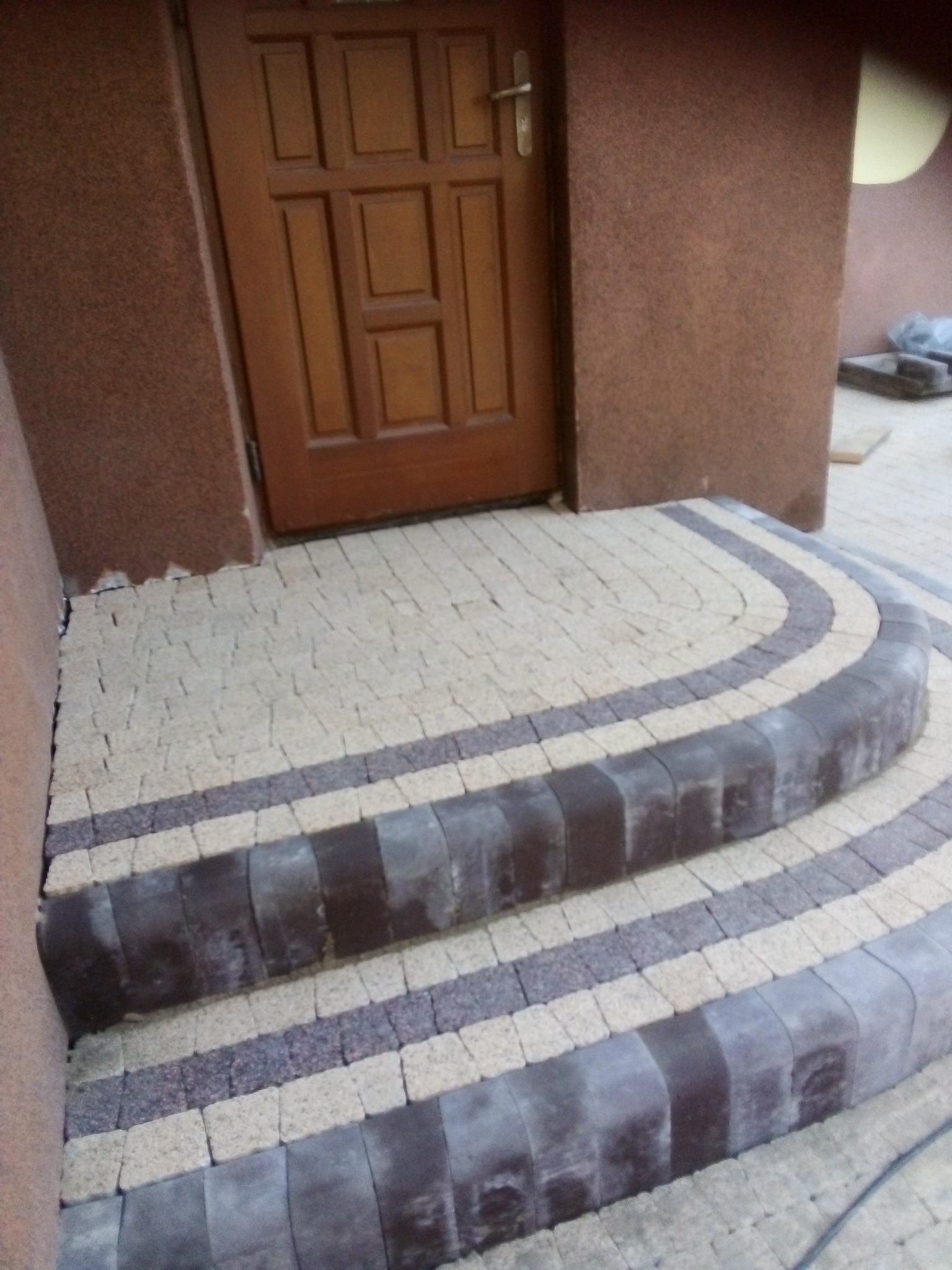 Schody wejściowe wyłożone kostką brukową w odcieniach beżu i grafitu, prowadzące do brązowych drzwi wejściowych.