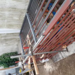 Konstrukcja metalowa ogrodzenia w trakcie spawania, zabezpieczona ściskami stolarskimi, w tle widoczny fragment roślinności i podłoże z betonu.