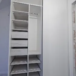 Wnętrze białej szafy przesuwnej z szufladami i półkami z jasnego drewna, widoczne metalowe prowadnice szuflad i drążek na ubrania.