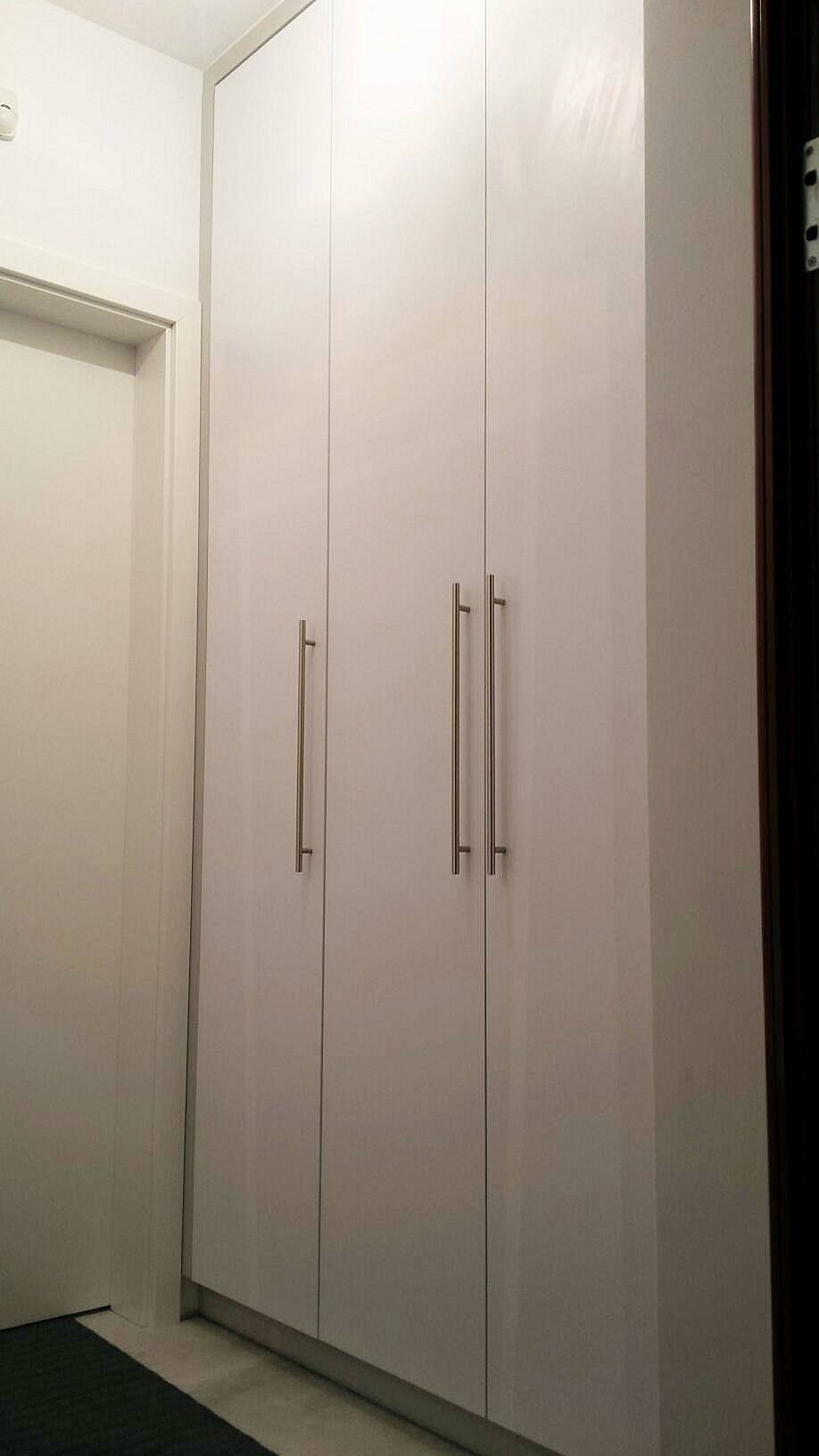 Wysoka, biała szafa wnękowa z czterema drzwiami uchylnymi i długimi, pionowymi uchwytami ze stali nierdzewnej, zamontowana w wąskim korytarzu.