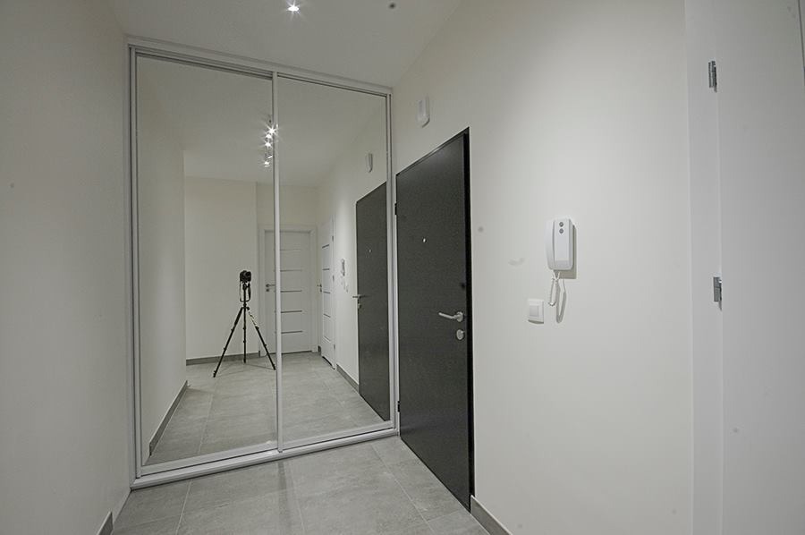 Korytarz z zabudowaną szafą z lustrzanymi drzwiami przesuwnymi, w której odbija się aparat na statywie.