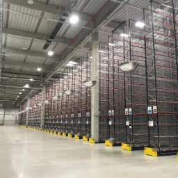 Wysokie, puste, metalowe regały magazynowe w perspektywie, oświetlone lampami sufitowymi, z żółto-czarnymi barierami ochronnymi przy słupach.