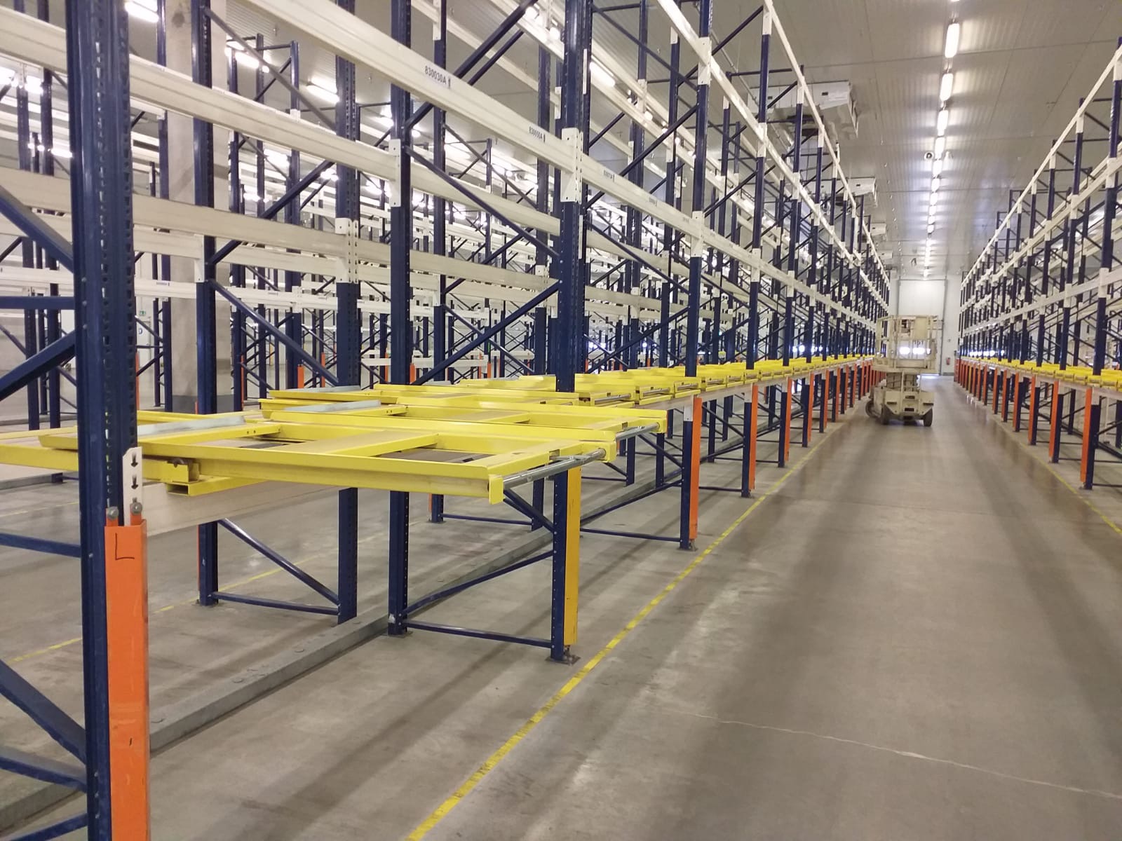 Długi korytarz magazynowy z rzędami metalowych regałów w kolorze granatowym i żółtym, widoczny wózek widłowy na końcu korytarza.