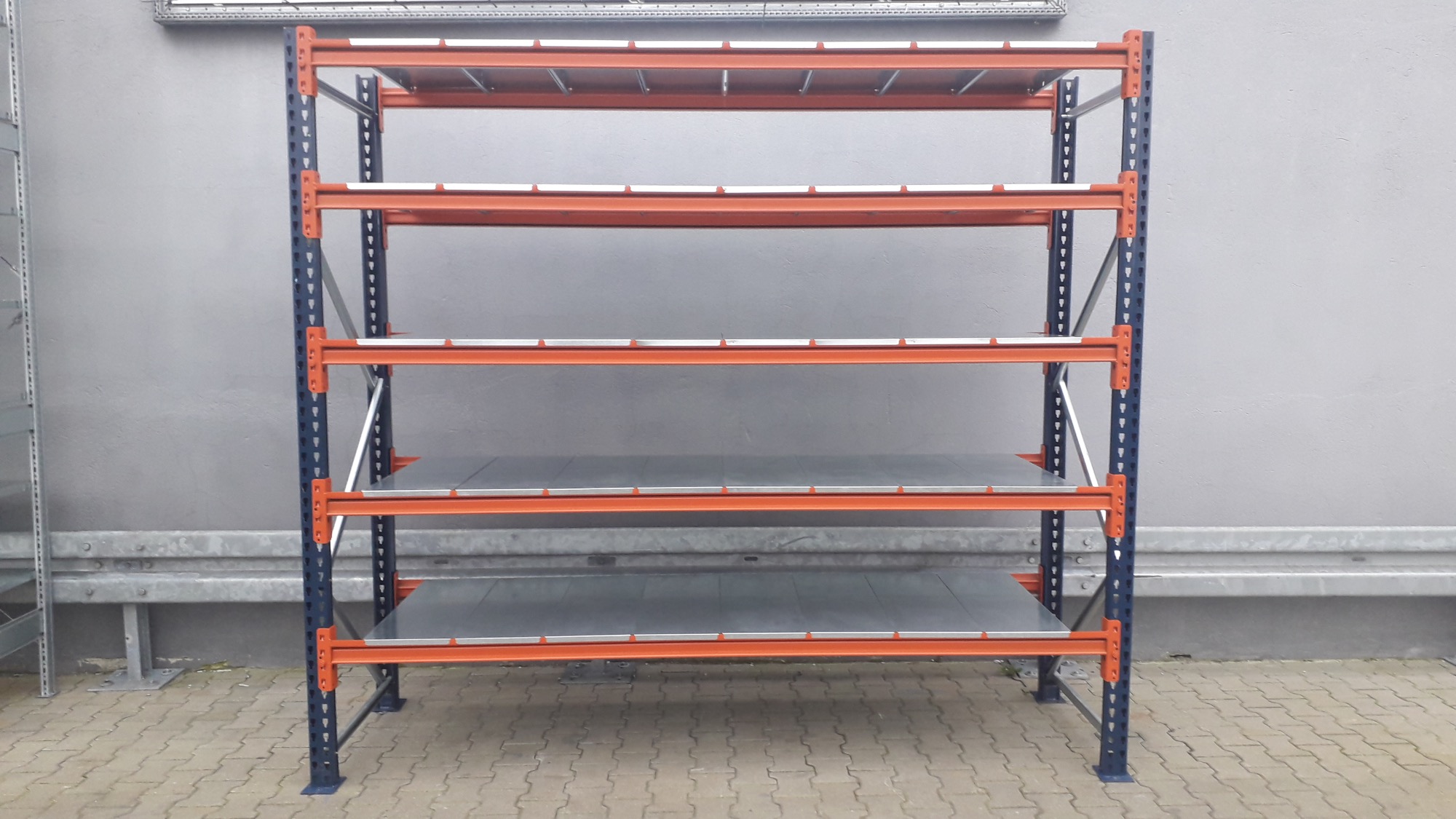 Metalowy regał magazynowy z czterema poziomami, pomarańczowo-granatowa konstrukcja, widok z przodu na tle szarej ściany i kostki brukowej.