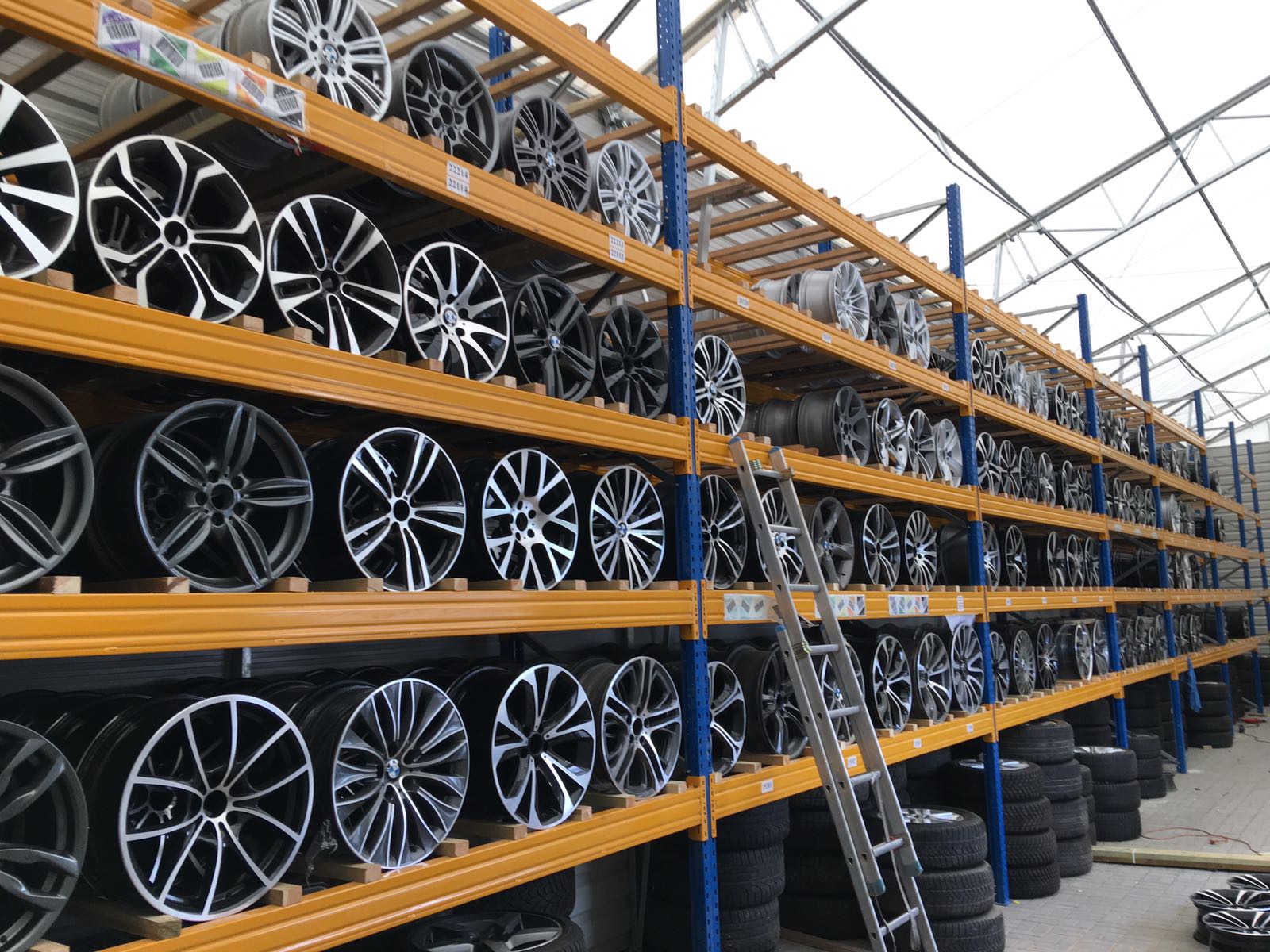 Wysokie, metalowe regały w magazynie, wypełnione różnymi wzorami felg samochodowych, z widoczną drabiną opartą o regał.