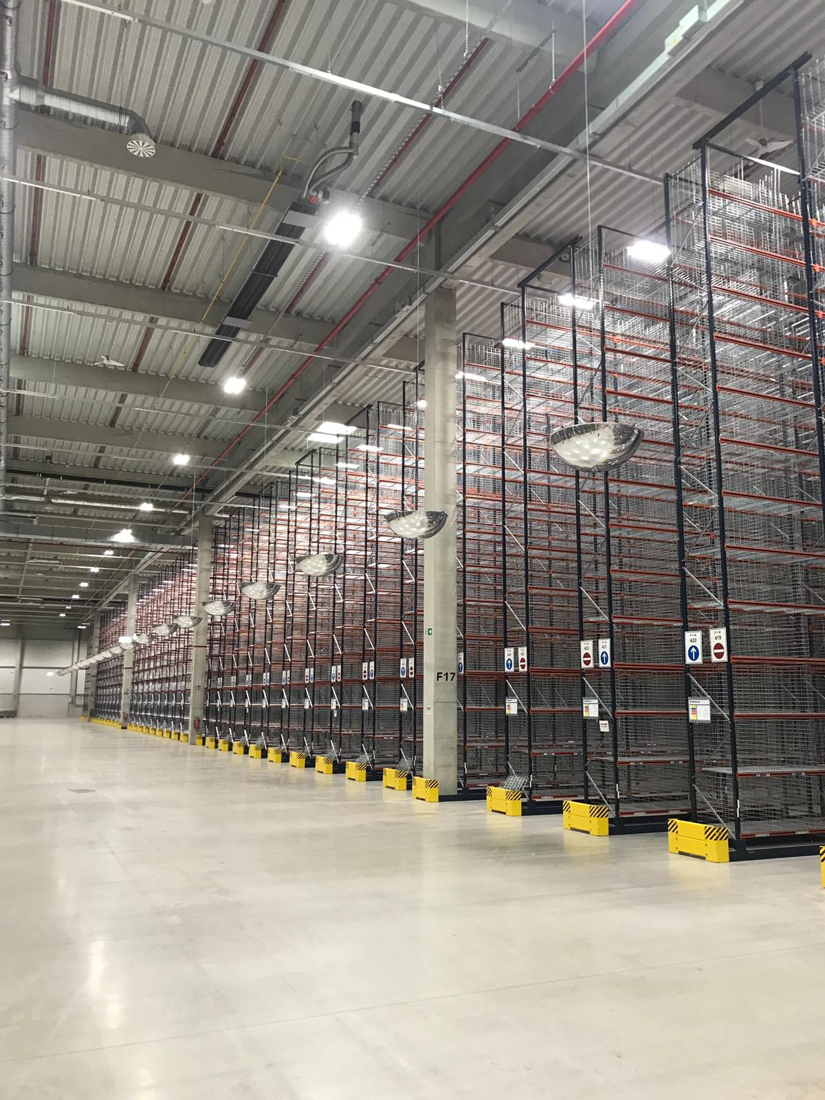 Wysokie, puste, metalowe regały magazynowe w perspektywie, oświetlone lampami sufitowymi, z żółto-czarnymi barierami ochronnymi przy słupach.