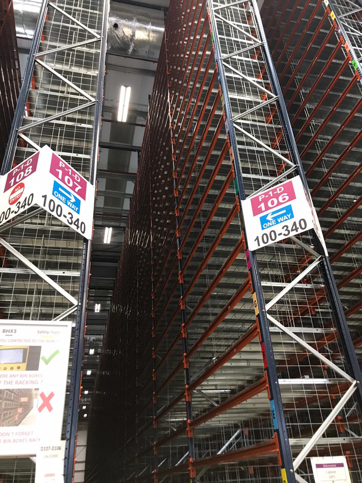 Wysokie, puste regały magazynowe z oznaczeniami nawigacyjnymi 'P-1-D 108' i 'P-1-D 106', widok z dołu w kierunku sufitu magazynu.