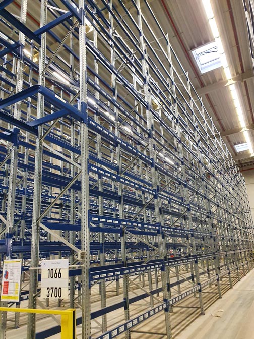 Wysokie, metalowe regały magazynowe w kolorze niebiesko-srebrnym, z widocznymi numerami identyfikacyjnymi 1066 i 3700, oświetlone światłem dziennym z okien dachowych.