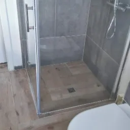 Nowoczesna kabina prysznicowa z szarymi płytkami ściennymi i drewnopodobną podłogą, widoczny fragment toalety, minimalistyczny design.