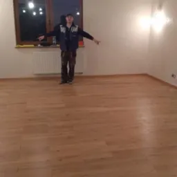 Jasne, nowo położone parkiety w pustym pokoju z białymi ścianami i oświetleniem halogenowym. Mężczyzna w roboczym ubraniu stoi na środku pomieszczenia, wskazując na podłogę.