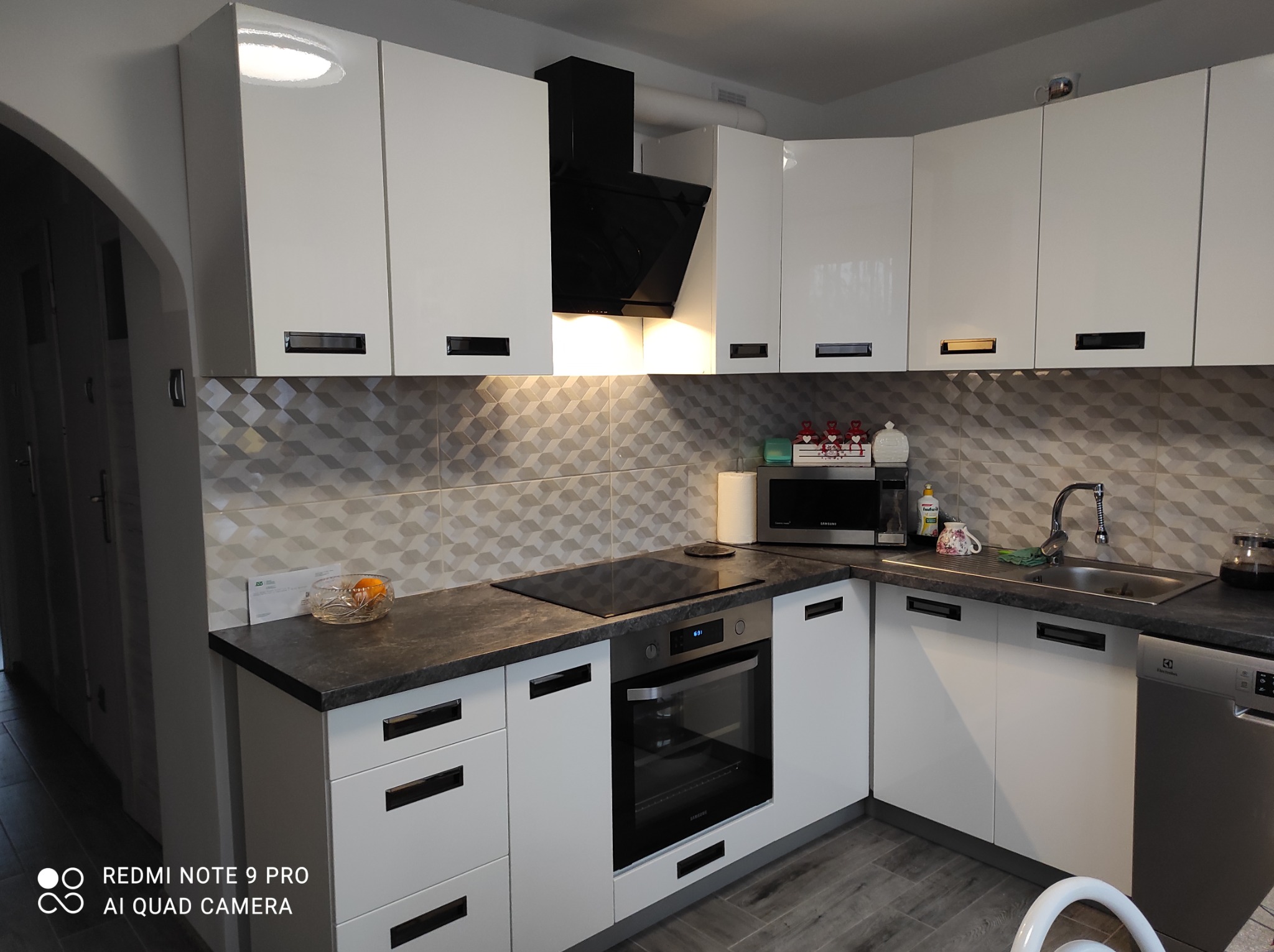 Nowoczesna kuchnia w bieli z czarnymi akcentami: szafki z połyskiem, czarny okap nad płytą indukcyjną, szara mozaika na ścianie, i elementy wyposażenia AGD.