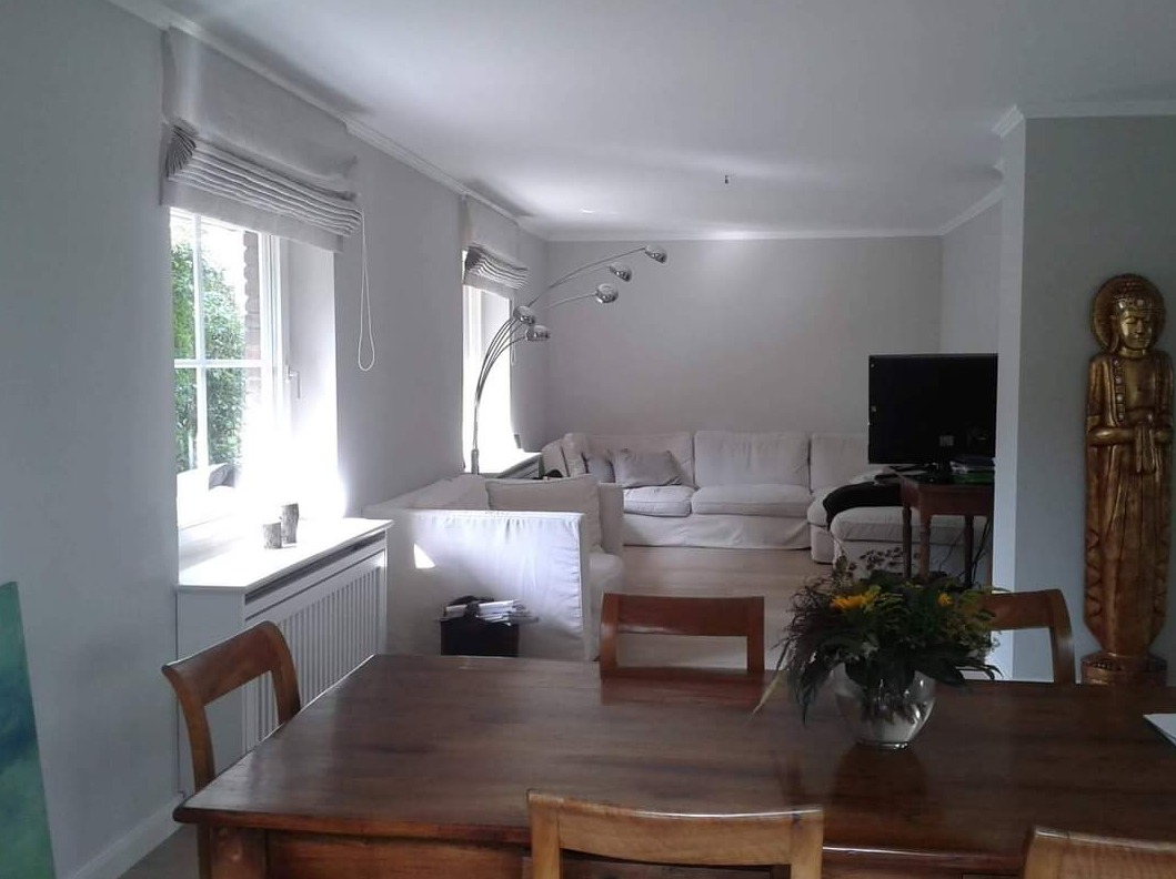 Jasne wnętrze salonu z drewnianym stołem na pierwszym planie, białymi roletami rzymskimi na oknach i rzeźbą Buddy. W tle biała sofa i lampa łukowa.