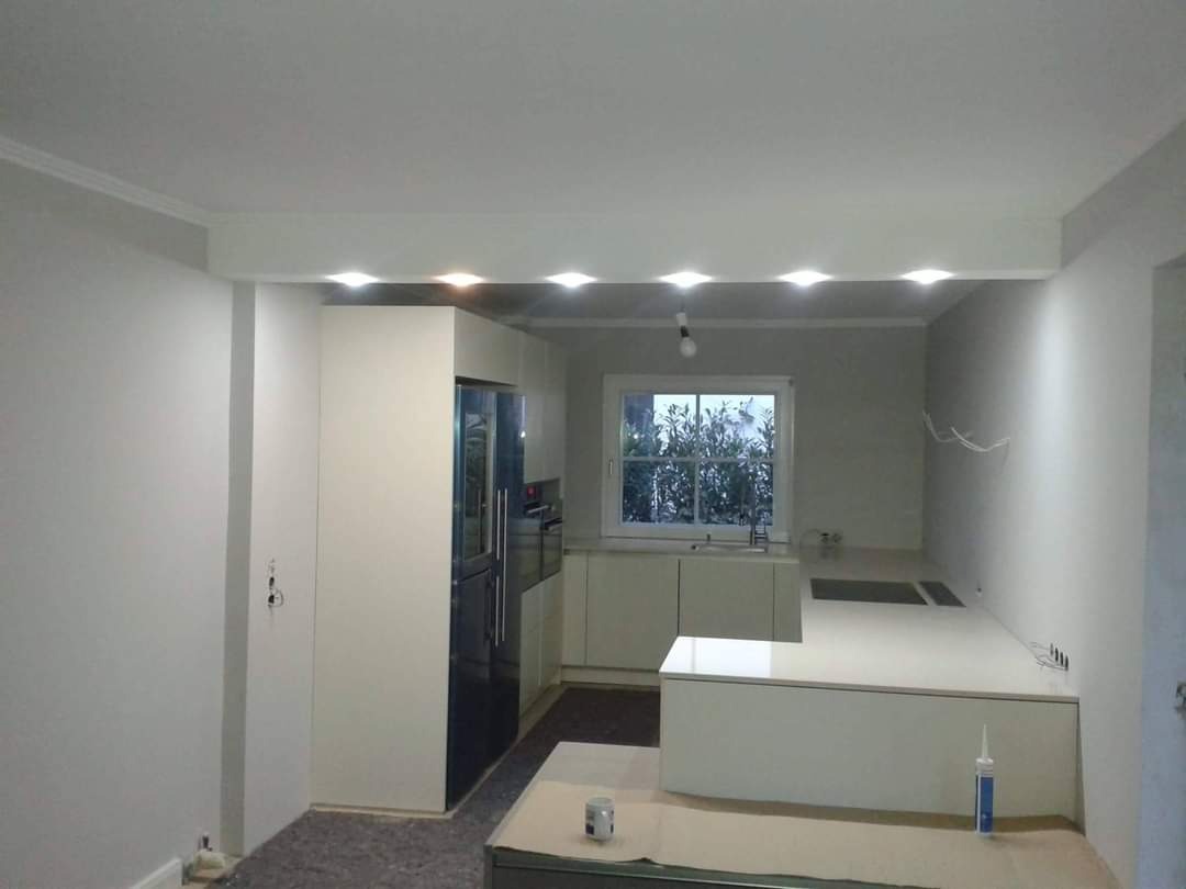 Wnętrze kuchni w trakcie remontu, widoczne białe szafki, blat roboczy, lodówka w zabudowie, podwieszany sufit z oświetleniem punktowym oraz niedokończone ściany i przewody elektryczne.