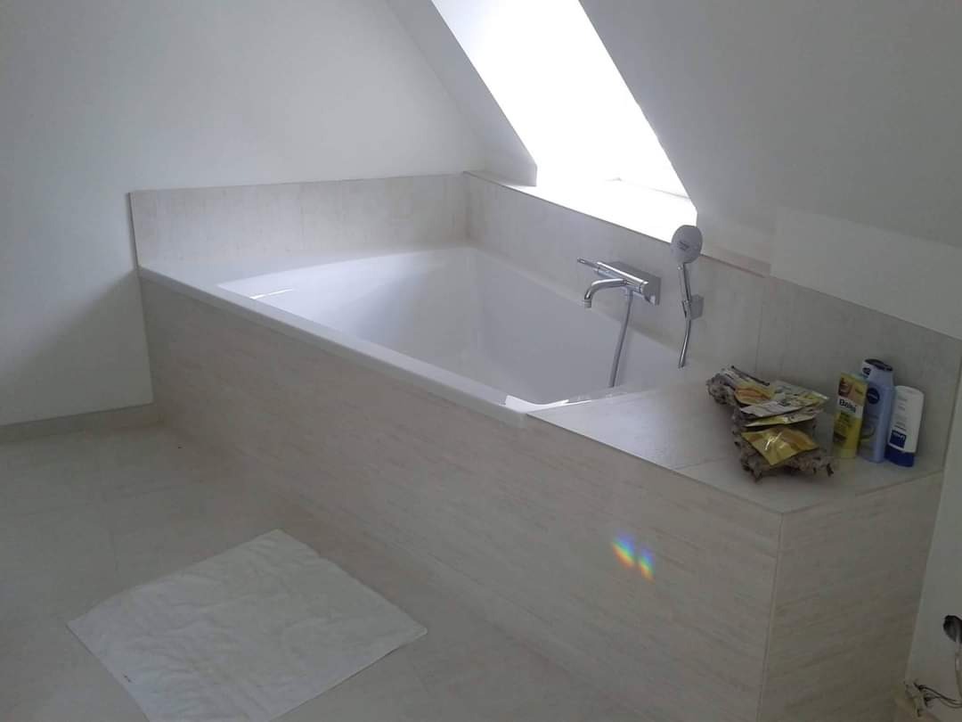 Biała wanna obudowana płytkami w łazience na poddaszu, widoczne kosmetyki i ręcznik na podłodze.