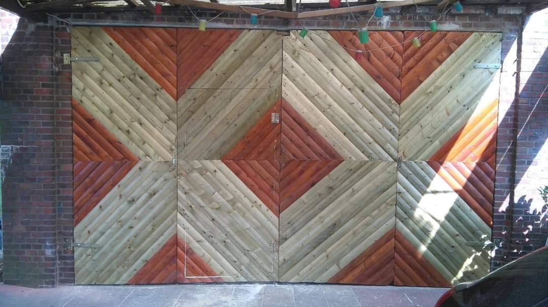 Czteroskrzydłowe, składane drzwi garażowe z drewna, malowane w ukośny wzór w odcieniach brązu i naturalnego drewna, zamontowane na ceglanym murze pod zadaszeniem z dekoracyjnymi lampkami.