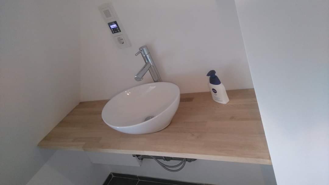 Owalna umywalka nablatowa z chromowaną baterią na drewnianym blacie w narożnej, minimalistycznej łazience z czarnymi płytkami podłogowymi i dozownikiem mydła Nivea obok.