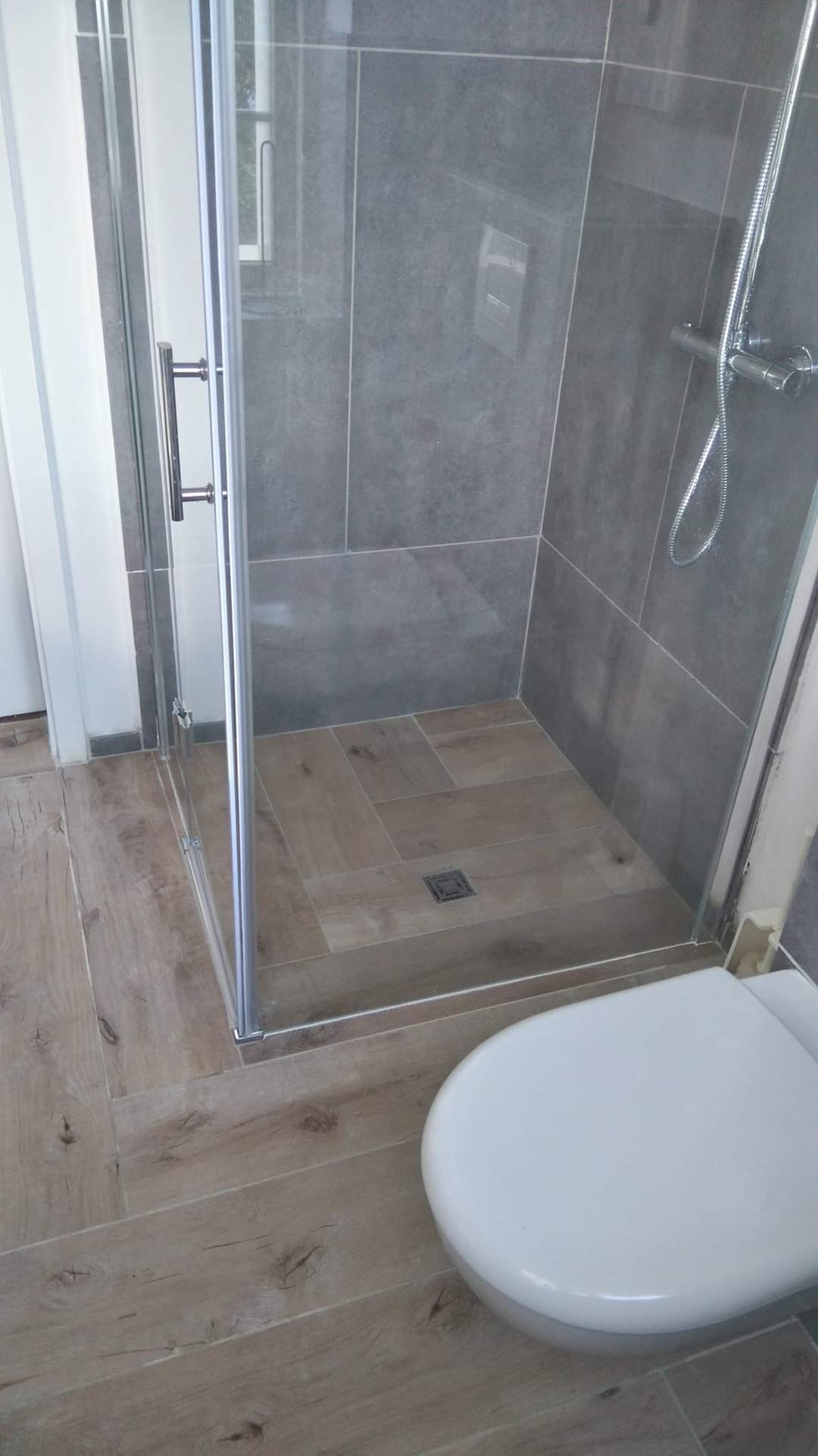 Nowoczesna kabina prysznicowa z szarymi płytkami ściennymi i drewnopodobną podłogą, widoczny fragment toalety, minimalistyczny design.