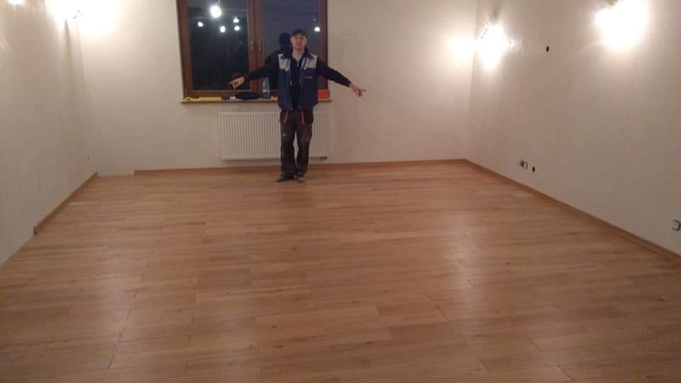 Jasne, nowo położone parkiety w pustym pokoju z białymi ścianami i oświetleniem halogenowym. Mężczyzna w roboczym ubraniu stoi na środku pomieszczenia, wskazując na podłogę.