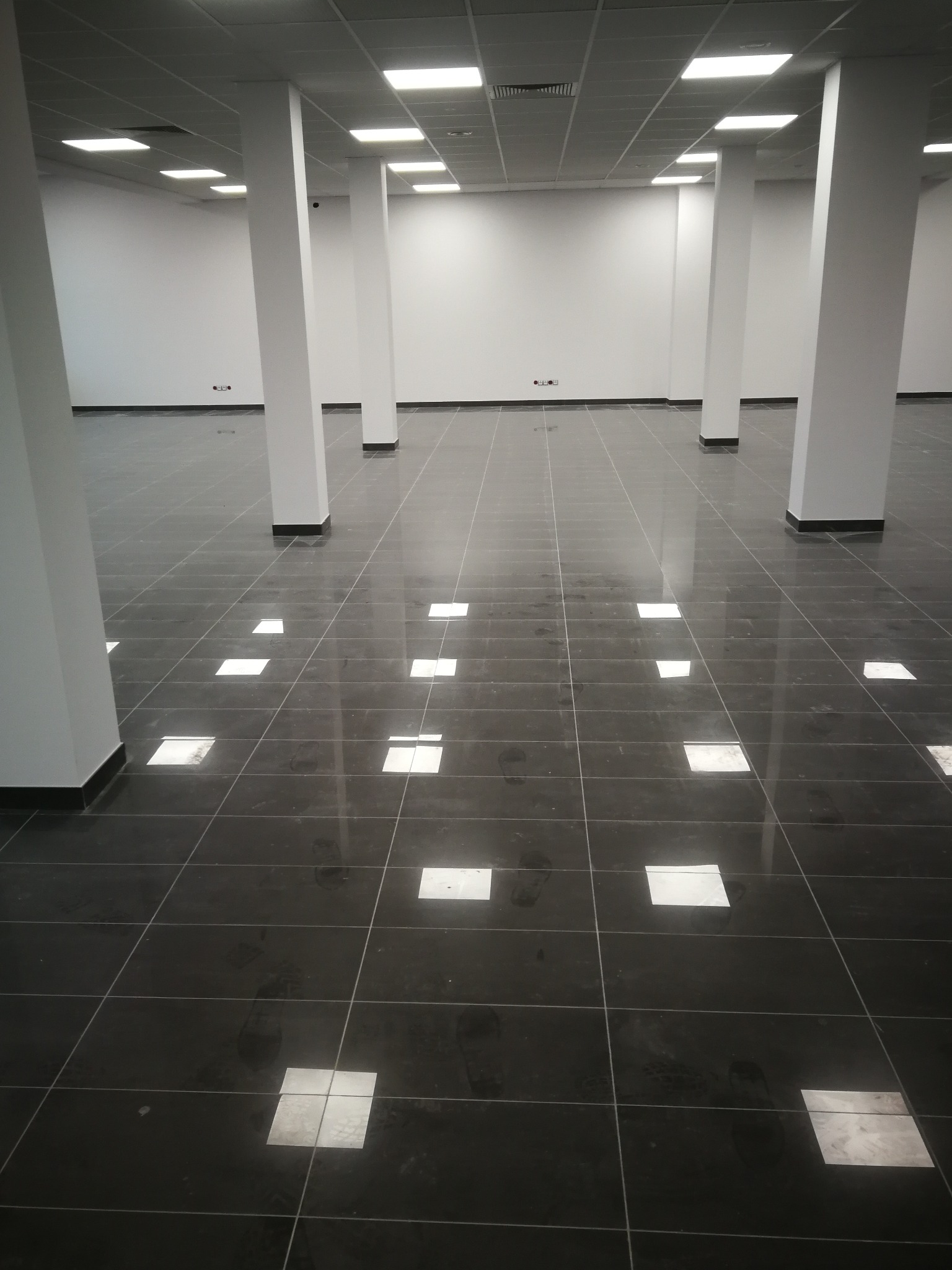 Wykończone wnętrze z czarną, błyszczącą podłogą z płytek ceramicznych, odbijającą światła sufitowe. Widoczne białe, kwadratowe kolumny i białe ściany. Na podłodze widoczne ślady butów.