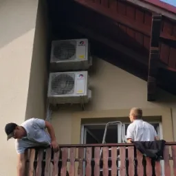 Dwaj monterzy na balkonie instalują dwie jednostki zewnętrzne klimatyzacji marki Gree na elewacji budynku, widoczna drabina oparta o okno.