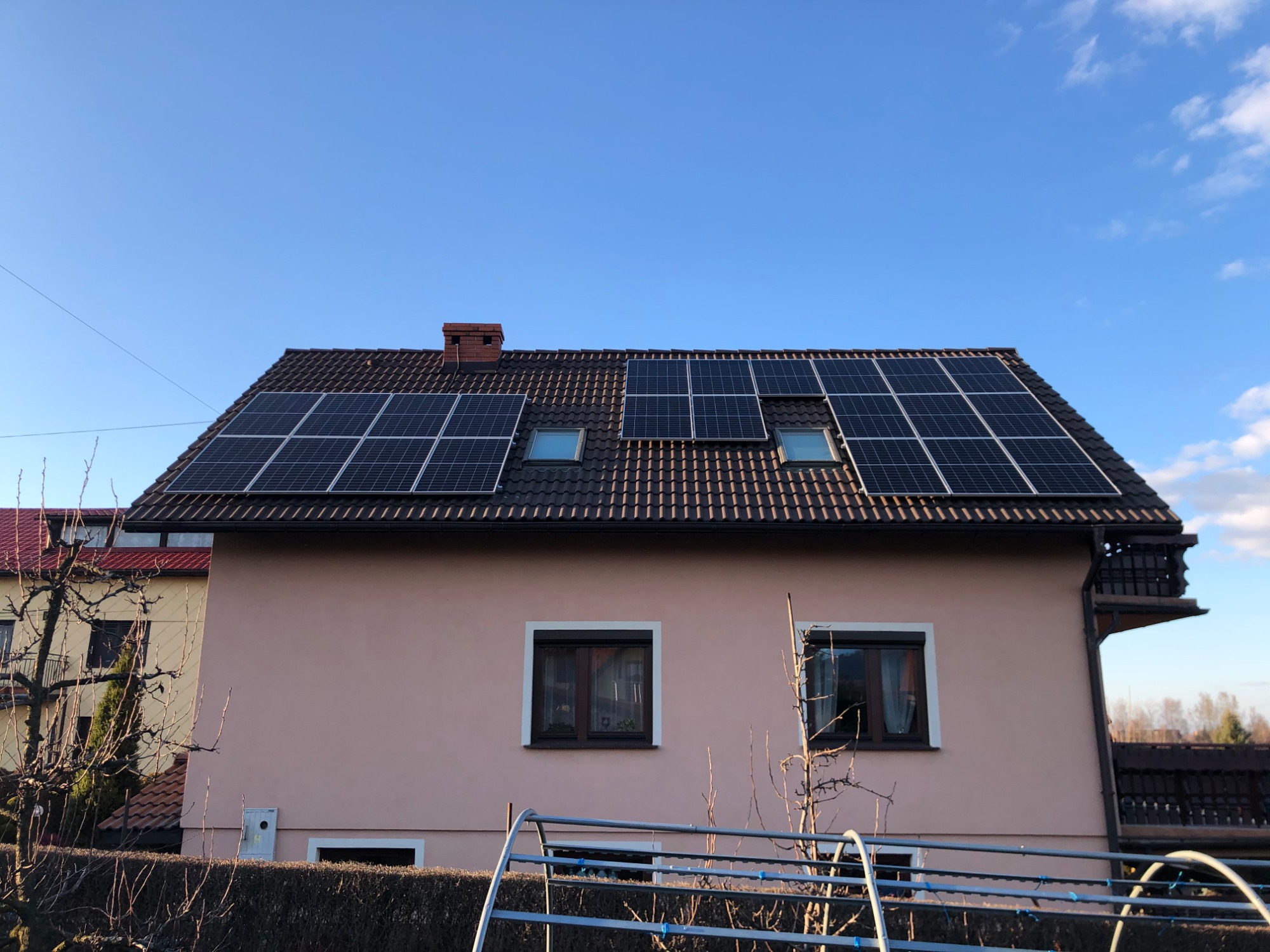 Dom jednorodzinny z brązowym dachem pokrytym panelami fotowoltaicznymi i dwoma oknami dachowymi, jasnoróżowa elewacja, błękitne niebo w tle.