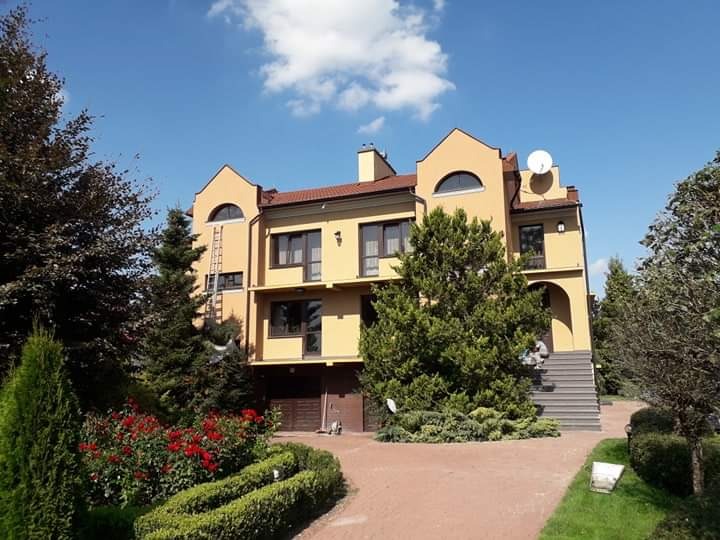 Żółty, dwupiętrowy dom z brązowymi oknami i dachem, otoczony zielenią, z drabiną opartą o ścianę i osobą siedzącą na schodach wejściowych.