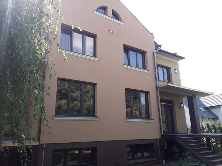 Elewacja dwukondygnacyjnego domu w Zamościu, ocieplona i pomalowana na beżowo, z ciemnymi oknami i brązowym cokołem, widok z boku, z fragmentem drzewa w lewym rogu.