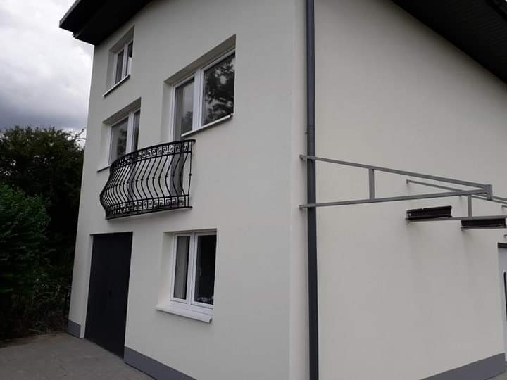 Jasna elewacja dwupiętrowego domu z czarnym balkonem i szarą podmurówką, widoczna rynna i zadaszenie.