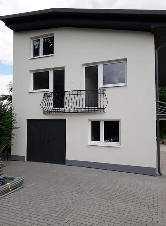 Dwupiętrowy budynek z szarą elewacją i czarnymi detalami, takimi jak dach, brama garażowa i balustrada balkonowa, widziany z poziomu kostki brukowej, z lekkim pochyleniem w lewo i stertą rusztowań...