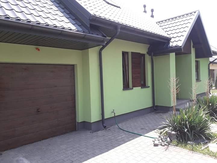 Dom jednorodzinny z zieloną elewacją, brązową bramą garażową i ciemnymi rynnami, widok z poziomu chodnika z kostki brukowej, z zielonym wężem ogrodowym przy ścianie.