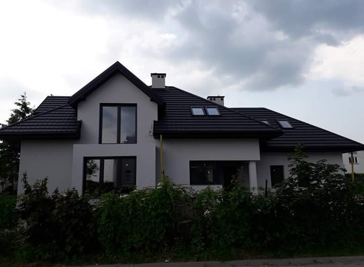 Nowoczesny dom jednorodzinny z szarą elewacją i czarnym dachem z dachówki, widoczne okna dachowe i komin, otoczony zielenią w pochmurny dzień.