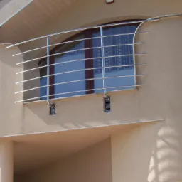 Zewnętrzna balustrada balkonowa z rurek ze stali nierdzewnej, zamocowana do elewacji budynku o jasnobeżowej, chropowatej strukturze tynku. Widoczne uchwyty mocujące balustradę do ściany. W tle okno...