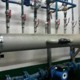 Poziomy, stalowy kolektor z licznymi zaworami i rurami, zamontowany w kotłowni, widoczne połączenia spawane i elementy mocujące.