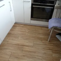 Jasne, drewnopodobne płytki podłogowe ułożone w kuchni z białymi szafkami i piekarnikiem, krzesło z poduszką w rogu.