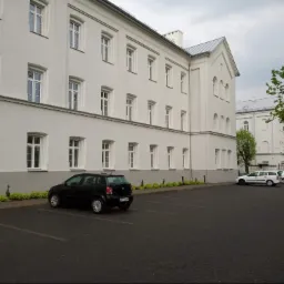 Odnowiony budynek z białymi ścianami, parkingiem z kostki brukowej i starannie przyciętymi krzewami, widoczne dwa zaparkowane samochody osobowe.