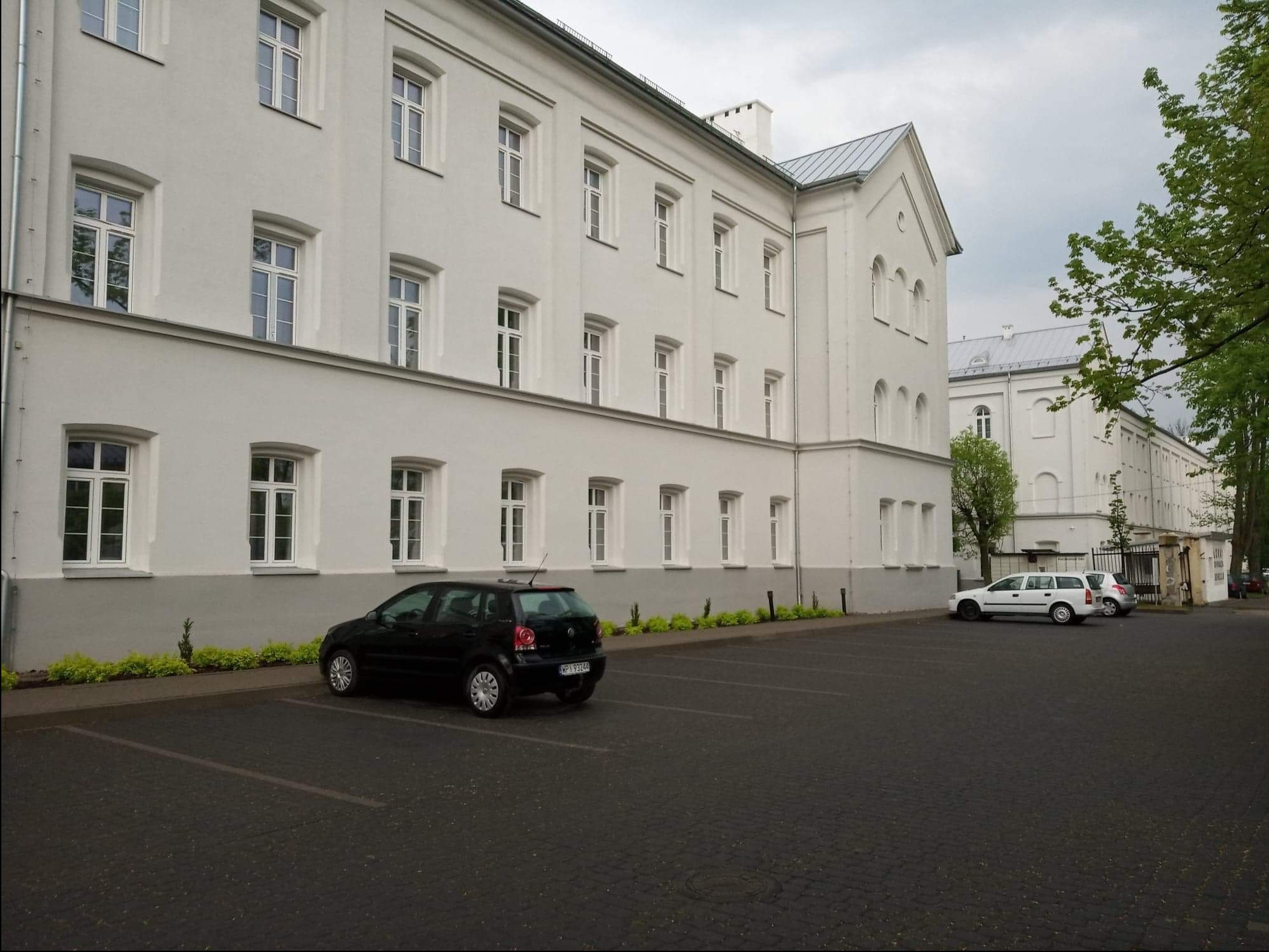Odnowiony budynek z białymi ścianami, parkingiem z kostki brukowej i starannie przyciętymi krzewami, widoczne dwa zaparkowane samochody osobowe.