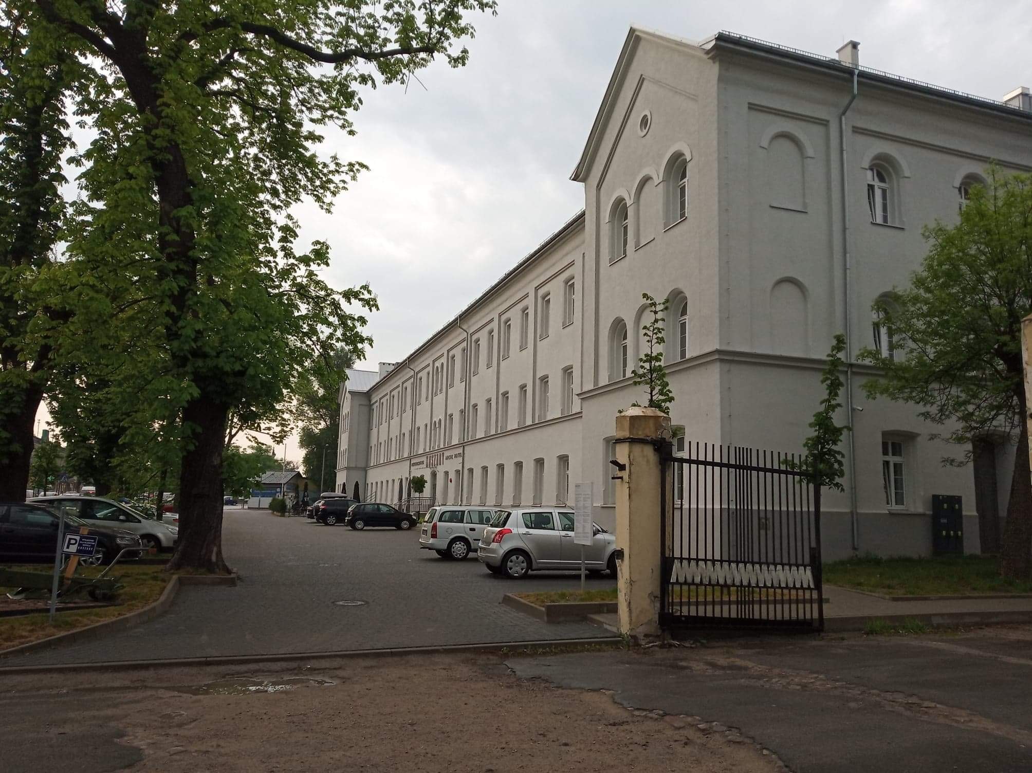 Długi, jasny budynek biurowy w stylu klasycystycznym z rzędami okien, widoczny zza metalowej bramy i parkingu z samochodami.