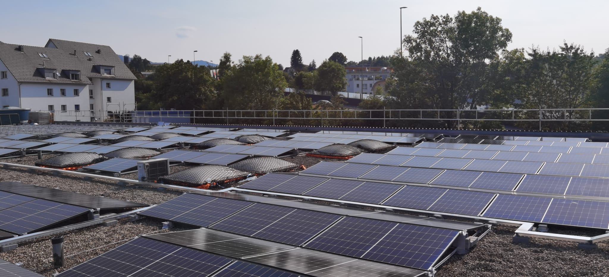 Rozległa instalacja fotowoltaiczna na dachu budynku z widocznymi panelami, systemem montażowym i elementami zabezpieczającymi. W tle zabudowa miejska i zieleń.