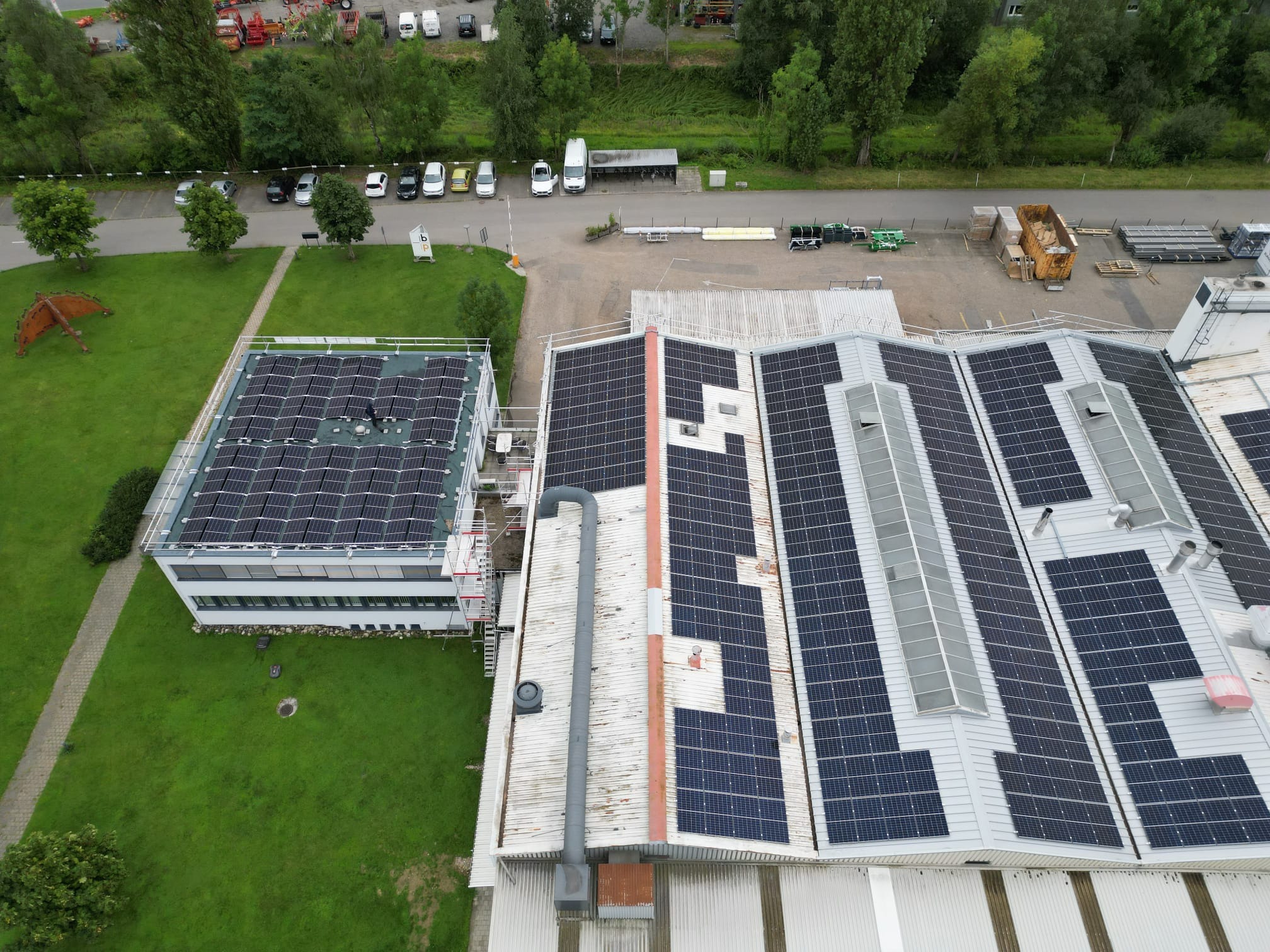 Widok z góry na budynki z panelami słonecznymi na dachu. Panele różnią się układem i wielkością. Na trawniku rzeźba. W tle parking z samochodami i zieleń.
