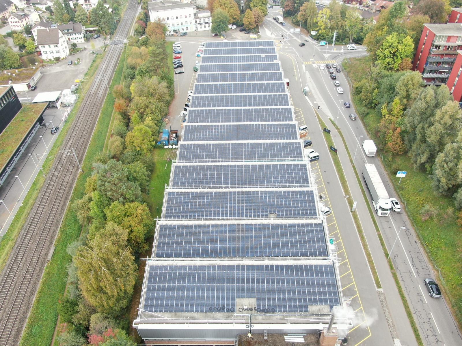 Rozległa instalacja fotowoltaiczna na dachu budynku, widok z góry, z linią kolejową i ulicą w tle. Uwagę zwraca dym wydobywający się z komina.