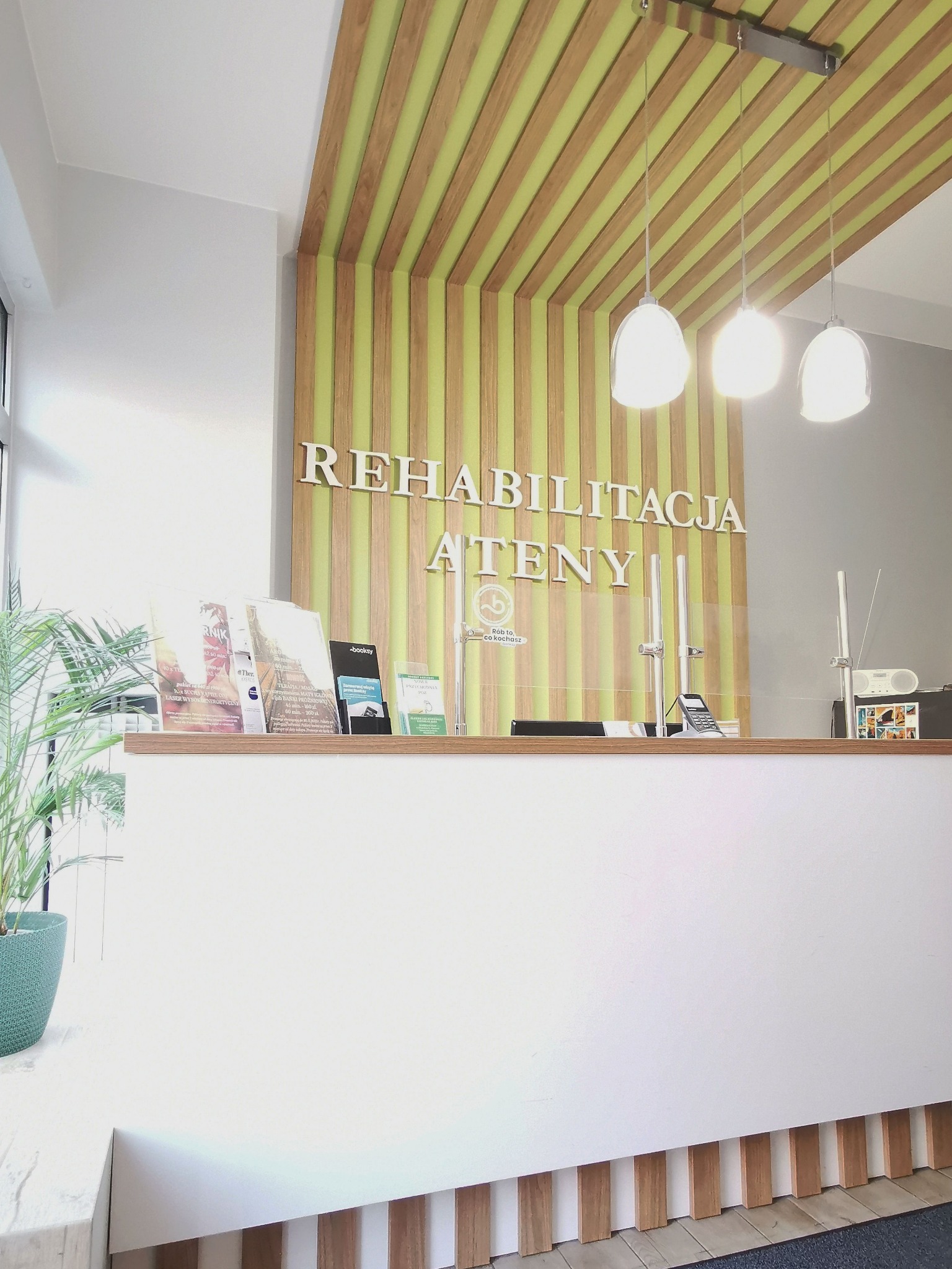 Recepcja w jasnych barwach z drewnianymi elementami dekoracyjnymi na suficie i ścianie, widoczne materiały informacyjne i logo 'Rehabilitacja Ateny'.